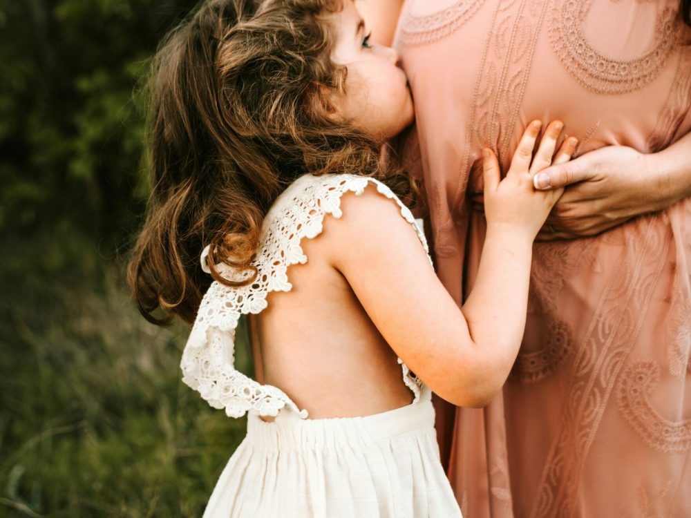 girl in white dress hugging pregnant mother wearing pink dress in nature